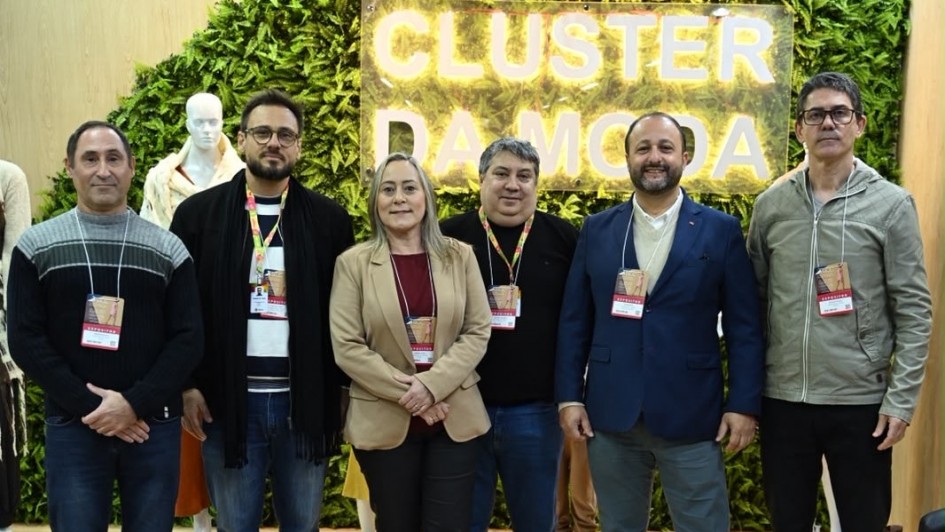 Seis pessoas posam para a foto em frente a um painel verde com as palavras "Cluster da Moda" em destaque. 