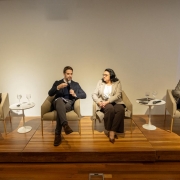 Vis&atilde;o frontal do painel com quatro participantes sentados lado a lado em poltronas sobre o palco, com pequenas mesas de apoio entre eles e proje&ccedil;&otilde;es laterais com elementos gr&aacute;ficos e as palavras &ldquo;Novas Energias&rdquo;.