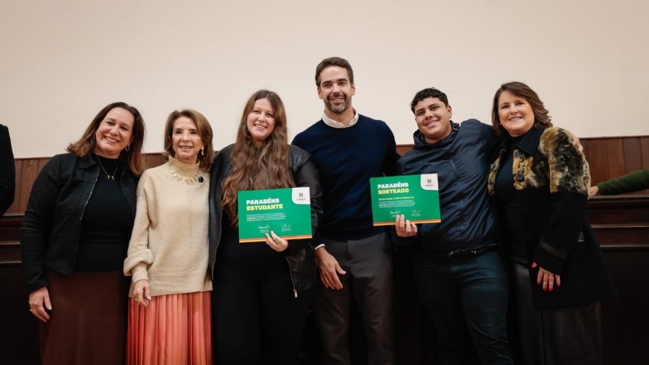 Aqui est&aacute; a descri&ccedil;&atilde;o da imagem para fins de acessibilidade (texto alternativo / #PraCegoVer):

---

**Descri&ccedil;&atilde;o da imagem "Governo do Estado realiza ato de celebra&ccedil;&atilde;o de estudantes destaque do Saers 2025.jpeg":**

Uma fotografia colorida em plano m&eacute;dio e vis&atilde;o frontal mostra um grupo de seis pessoas sorrindo lado a lado em um ambiente interno. Elas est&atilde;o posicionadas &agrave; frente de uma parede lisa de tom claro (bege) na metade superior e com revestimento de madeira escura na metade inferior.

**Participantes (da esquerda para a direita):**

1. Uma mulher de cabelos castanhos longos, vestindo uma jaqueta preta aberta sobre uma blusa preta, colar dourado fino e uma saia longa em tom marrom-escuro.
2. Uma mulher de cabelos castanhos curtos e ondulados, usando uma blusa de tric&ocirc; de gola alta bege-clara, colar dourado e uma saia longa plissada em tom bronze ou salm&atilde;o-escuro.
3. Uma jovem de cabelos castanhos bem longos, vestindo uma jaqueta preta de material sint&eacute;tico sobre roupa preta. Ela segura com as duas m&atilde;os um certificado verde com bordas inferiores coloridas, onde se l&ecirc; em destaque "PARAB&Eacute;NS ESTUDANTE".
4. Um homem (Eduardo Leite) de barba curta aparada, vestindo um su&eacute;ter azul-marinho sobre camisa branca com gola aparecendo e cal&ccedil;a de alfaiataria cinza-escura. Ele est&aacute; posicionado no centro do grupo, com o bra&ccedil;o discretamente atr&aacute;s da jovem &agrave; sua esquerda.
5. Um rapaz jovem de cabelos pretos cacheados e curtos, vestindo uma jaqueta esportiva azul-escura fechada e cal&ccedil;a escura. Ele sorri e segura com a m&atilde;o esquerda um certificado verde id&ecirc;ntico ao da colega, onde se l&ecirc; "PARAB&Eacute;NS SORTEADO". O bra&ccedil;o direito de outra pessoa repousa sobre seu ombro.
6. Uma mulher de cabelos castanhos na altura dos ombros, usando um casaco preto comprido com detalhes em estampa de pele de animal (leopardo/on&ccedil;a) na gola e nos ombros, sobre blusa preta, e cal&ccedil;a escura. Ela est&aacute; com o bra&ccedil;o esquerdo estendido atr&aacute;s do rapaz ao seu lado.

**Ilumina&ccedil;&atilde;o e enquadramento:**
A ilumina&ccedil;&atilde;o &eacute; frontal e uniforme, destacando os rostos e os certificados verdes decorados com as cores da bandeira do estado (rio-grandense) na borda inferior. O grupo est&aacute; ligeiramente inclinado de forma descontra&iacute;da uns em dire&ccedil;&atilde;o aos outros, transmitindo uma atmosfera de celebra&ccedil;&atilde;o e conquista.