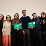 Aqui est&aacute; a descri&ccedil;&atilde;o da imagem para fins de acessibilidade (texto alternativo / #PraCegoVer):

---

**Descri&ccedil;&atilde;o da imagem "Governo do Estado realiza ato de celebra&ccedil;&atilde;o de estudantes destaque do Saers 2025.jpeg":**

Uma fotografia colorida em plano m&eacute;dio e vis&atilde;o frontal mostra um grupo de seis pessoas sorrindo lado a lado em um ambiente interno. Elas est&atilde;o posicionadas &agrave; frente de uma parede lisa de tom claro (bege) na metade superior e com revestimento de madeira escura na metade inferior.

**Participantes (da esquerda para a direita):**

1. Uma mulher de cabelos castanhos longos, vestindo uma jaqueta preta aberta sobre uma blusa preta, colar dourado fino e uma saia longa em tom marrom-escuro.
2. Uma mulher de cabelos castanhos curtos e ondulados, usando uma blusa de tric&ocirc; de gola alta bege-clara, colar dourado e uma saia longa plissada em tom bronze ou salm&atilde;o-escuro.
3. Uma jovem de cabelos castanhos bem longos, vestindo uma jaqueta preta de material sint&eacute;tico sobre roupa preta. Ela segura com as duas m&atilde;os um certificado verde com bordas inferiores coloridas, onde se l&ecirc; em destaque "PARAB&Eacute;NS ESTUDANTE".
4. Um homem (Eduardo Leite) de barba curta aparada, vestindo um su&eacute;ter azul-marinho sobre camisa branca com gola aparecendo e cal&ccedil;a de alfaiataria cinza-escura. Ele est&aacute; posicionado no centro do grupo, com o bra&ccedil;o discretamente atr&aacute;s da jovem &agrave; sua esquerda.
5. Um rapaz jovem de cabelos pretos cacheados e curtos, vestindo uma jaqueta esportiva azul-escura fechada e cal&ccedil;a escura. Ele sorri e segura com a m&atilde;o esquerda um certificado verde id&ecirc;ntico ao da colega, onde se l&ecirc; "PARAB&Eacute;NS SORTEADO". O bra&ccedil;o direito de outra pessoa repousa sobre seu ombro.
6. Uma mulher de cabelos castanhos na altura dos ombros, usando um casaco preto comprido com detalhes em estampa de pele de animal (leopardo/on&ccedil;a) na gola e nos ombros, sobre blusa preta, e cal&ccedil;a escura. Ela est&aacute; com o bra&ccedil;o esquerdo estendido atr&aacute;s do rapaz ao seu lado.

**Ilumina&ccedil;&atilde;o e enquadramento:**
A ilumina&ccedil;&atilde;o &eacute; frontal e uniforme, destacando os rostos e os certificados verdes decorados com as cores da bandeira do estado (rio-grandense) na borda inferior. O grupo est&aacute; ligeiramente inclinado de forma descontra&iacute;da uns em dire&ccedil;&atilde;o aos outros, transmitindo uma atmosfera de celebra&ccedil;&atilde;o e conquista.