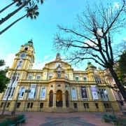  Imagem mostra um grande edif&iacute;cio hist&oacute;rico de arquitetura ecl&eacute;tica/neocl&aacute;ssica, fotografado de um &acirc;ngulo baixo e com lente grande-angular, o que destaca sua impon&ecirc;ncia e altura. No centro da cena est&aacute; uma constru&ccedil;&atilde;o de fachada predominantemente amarela e bege, com elementos ornamentais em branco. O edif&iacute;cio possui uma composi&ccedil;&atilde;o sim&eacute;trica, com uma entrada principal formada por tr&ecirc;s grandes arcos na parte t&eacute;rrea. 