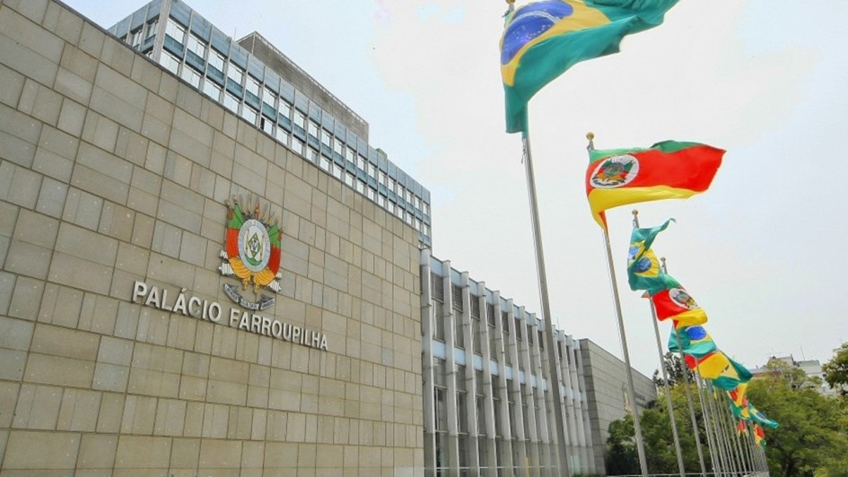Aqui est&aacute; a descri&ccedil;&atilde;o da imagem para fins de acessibilidade (texto alternativo / #PraCegoVer):

---

**Descri&ccedil;&atilde;o da imagem "Assembleia aprova contrata&ccedil;&atilde;o de empr&eacute;stimo externo pelo Estado para enfrentamento de desastres naturais.jpg":**

Uma fotografia colorida em plano geral e &acirc;ngulo contra-plong&eacute;e (de baixo para cima) mostra a fachada externa do Pal&aacute;cio Farroupilha, sede da Assembleia Legislativa do Estado do Rio Grande do Sul, sob um c&eacute;u claro e ligeiramente encoberto.

**O Edif&iacute;cio:**

* &Agrave; esquerda, destaca-se uma grande parede de blocos de pedra retangulares em tom bege-claro. Fixado no centro dessa parede, h&aacute; um bras&atilde;o de armas colorido e tridimensional e, logo abaixo dele, o nome **"PAL&Aacute;CIO FARROUPILHA"** em letras met&aacute;licas prateadas e em caixa alta.
* Ao centro e estendendo-se para a direita, a arquitetura do pr&eacute;dio continua com uma estrutura moderna composta por grandes janelas de vidro verticais intercaladas por pilares cinzas e azuis-claros. Uma parte mais alta do edif&iacute;cio, com m&uacute;ltiplas janelas, se projeta ao fundo.

**As Bandeiras:**
No canto superior direito, uma fileira de mastros met&aacute;licos se estende em perspectiva diagonal em dire&ccedil;&atilde;o ao fundo. V&aacute;rias bandeiras oficiais aparecem hasteadas e ondulando com o vento:

* A primeira bandeira, mais alta e em destaque, &eacute; a **bandeira do Brasil** (verde e amarela com o c&iacute;rculo azul central).
* A segunda bandeira &eacute; a **bandeira do estado do Rio Grande do Sul** (com suas listras diagonais em verde, vermelho e amarelo e o bras&atilde;o centralizado).
* Atr&aacute;s delas, o padr&atilde;o se repete com outras bandeiras do Brasil e do Estado hasteadas em sequ&ecirc;ncia ao longo da esplanada.

**Elementos Adicionais:**
Na parte inferior, em frente ao pr&eacute;dio, nota-se uma rampa ou passarela de acesso com guarda-corpo met&aacute;lico branco. No canto inferior direito, ao fundo, aparecem copas de &aacute;rvores verdes e uma pequena por&ccedil;&atilde;o da rua adjacente.