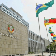 Aqui est&aacute; a descri&ccedil;&atilde;o da imagem para fins de acessibilidade (texto alternativo / #PraCegoVer):

---

**Descri&ccedil;&atilde;o da imagem "Assembleia aprova contrata&ccedil;&atilde;o de empr&eacute;stimo externo pelo Estado para enfrentamento de desastres naturais.jpg":**

Uma fotografia colorida em plano geral e &acirc;ngulo contra-plong&eacute;e (de baixo para cima) mostra a fachada externa do Pal&aacute;cio Farroupilha, sede da Assembleia Legislativa do Estado do Rio Grande do Sul, sob um c&eacute;u claro e ligeiramente encoberto.

**O Edif&iacute;cio:**

* &Agrave; esquerda, destaca-se uma grande parede de blocos de pedra retangulares em tom bege-claro. Fixado no centro dessa parede, h&aacute; um bras&atilde;o de armas colorido e tridimensional e, logo abaixo dele, o nome **"PAL&Aacute;CIO FARROUPILHA"** em letras met&aacute;licas prateadas e em caixa alta.
* Ao centro e estendendo-se para a direita, a arquitetura do pr&eacute;dio continua com uma estrutura moderna composta por grandes janelas de vidro verticais intercaladas por pilares cinzas e azuis-claros. Uma parte mais alta do edif&iacute;cio, com m&uacute;ltiplas janelas, se projeta ao fundo.

**As Bandeiras:**
No canto superior direito, uma fileira de mastros met&aacute;licos se estende em perspectiva diagonal em dire&ccedil;&atilde;o ao fundo. V&aacute;rias bandeiras oficiais aparecem hasteadas e ondulando com o vento:

* A primeira bandeira, mais alta e em destaque, &eacute; a **bandeira do Brasil** (verde e amarela com o c&iacute;rculo azul central).
* A segunda bandeira &eacute; a **bandeira do estado do Rio Grande do Sul** (com suas listras diagonais em verde, vermelho e amarelo e o bras&atilde;o centralizado).
* Atr&aacute;s delas, o padr&atilde;o se repete com outras bandeiras do Brasil e do Estado hasteadas em sequ&ecirc;ncia ao longo da esplanada.

**Elementos Adicionais:**
Na parte inferior, em frente ao pr&eacute;dio, nota-se uma rampa ou passarela de acesso com guarda-corpo met&aacute;lico branco. No canto inferior direito, ao fundo, aparecem copas de &aacute;rvores verdes e uma pequena por&ccedil;&atilde;o da rua adjacente.