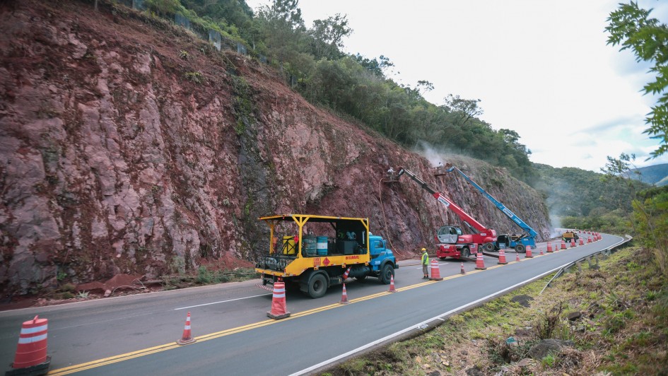 Trecho de rodovia em obra com cones sinalizando a pista, onde caminh&otilde;es e equipamentos operam junto a um pared&atilde;o rochoso; ao lado, um ve&iacute;culo de apoio com estrutura met&aacute;lica transporta materiais, enquanto trabalhadores atuam pr&oacute;ximos ao talude em meio &agrave; vegeta&ccedil;&atilde;o.