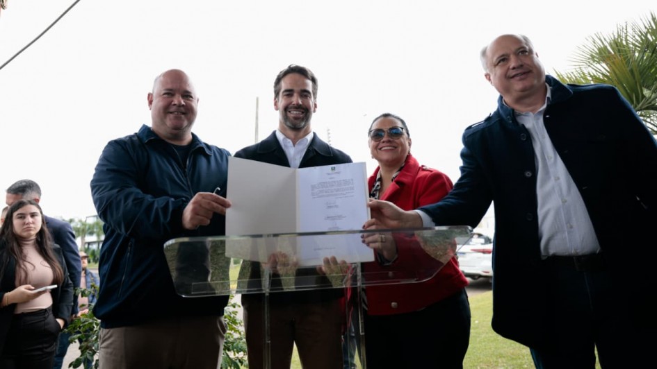 Momento de registro ap&oacute;s a assinatura, com quatro pessoas exibindo um documento aberto sobre o p&uacute;lpito para a c&acirc;mera, em um ambiente externo com gramado e participantes ao redor.