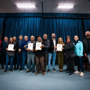 Aqui est&aacute; a descri&ccedil;&atilde;o da imagem para fins de acessibilidade 

---

**Descri&ccedil;&atilde;o da imagem "Governo do Estado entrega 42 t&iacute;tulos de legitima&ccedil;&atilde;o fundi&aacute;ria em Sapucaia do Sul e Gravata&iacute; por meio do Programa Regulariza RS.jpg":**

Uma fotografia colorida em plano geral e vis&atilde;o frontal mostra um grupo de cerca de 14 pessoas (homens e mulheres) posando alinhadas lado a lado em um palco ou espa&ccedil;o interno de eventos. Elas est&atilde;o sorrindo, e algumas delas seguram pastas brancas contendo documentos oficiais.

**Os Participantes e os Documentos:**

* Pelo menos quatro pessoas na linha de frente seguram de forma destacada pastas brancas verticais com o bras&atilde;o do Estado do Rio Grande do Sul e detalhes coloridos na borda inferior.
* As pessoas vestem roupas casuais de inverno e meia-esta&ccedil;&atilde;o, como jaquetas acolchoadas, casacos de moletom, su&eacute;teres tricotados, cal&ccedil;as jeans e t&ecirc;nis.
* No centro do grupo, destacam-se autoridades e cidad&atilde;os, incluindo um homem de cal&ccedil;a jeans clara e jaqueta preta aberta sobre camisa clara, uma mulher vestindo um macac&atilde;o ou conjunto verde-oliva formal, e uma mulher mais &agrave; direita com um casaco azul-turquesa de bot&otilde;es.

**O Cen&aacute;rio e Elementos de Fundo:**

* O grupo est&aacute; em p&eacute; sobre um piso liso na cor azul-claro acinzentado.
* Ao fundo, cobrindo toda a extens&atilde;o traseira, h&aacute; uma grande cortina de tecido brilhante em tom azul-petr&oacute;leo ou azul-marinho escuro, disposta em gomos verticais.
* No canto esquerdo, h&aacute; totens c&uacute;bicos brancos empilhados com a identidade visual e o logotipo do **"GOVERNO DO ESTADO RIO GRANDE DO SUL"**.
* No canto direito, h&aacute; uma pequena mesa coberta com uma toalha branca e, atr&aacute;s dela, parte de uma bandeira ou banner vertical onde se l&ecirc; parcialmente uma inscri&ccedil;&atilde;o em azul. O teto possui lumin&aacute;rias tubulares embutidas que clareiam o ambiente.