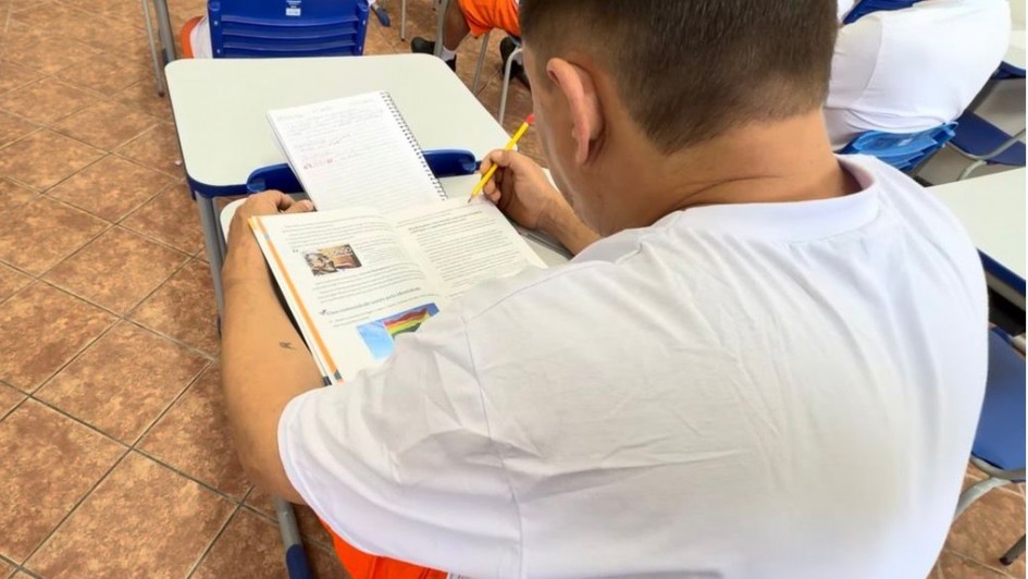 Aqui est&aacute; a descri&ccedil;&atilde;o da imagem para fins de acessibilidade 

---

**Descri&ccedil;&atilde;o da imagem "Sistema prisional do RS realiza mais de 12 mil atendimentos em educa&ccedil;&atilde;o formal e informal para pessoas privadas de liberdade.jpeg":**

Uma fotografia colorida em plano detalhe e &acirc;ngulo superior picado (visto de cima e por tr&aacute;s) foca em um detento sentado em sua carteira escolar durante uma atividade de estudo ou leitura em uma sala de aula.

**O Aluno e o Material Did&aacute;tico:**

* No centro e em primeiro plano, um homem de cabelos castanhos curt&iacute;ssimos e pele clara aparece de costas e de perfil parcial direito. Ele veste o uniforme padr&atilde;o do sistema prisional, composto por uma camiseta branca de manga curta e bermuda laranja-viva (parcialmente vis&iacute;vel na base). Ele segura um l&aacute;pis amarelo com a m&atilde;o direita, apontando para a p&aacute;gina, em uma postura concentrada de leitura. No antebra&ccedil;o esquerdo, nota-se uma pequena tatuagem minimalista escura.
* Sobre a mesa cinza com bordas azuis da carteira, h&aacute; um livro did&aacute;tico ou apostila com espiral aberto. Na p&aacute;gina esquerda do livro, h&aacute; textos impressos e duas ilustra&ccedil;&otilde;es coloridas: uma pequena imagem retangular na parte superior e, logo abaixo, a ilustra&ccedil;&atilde;o de uma bandeira com as cores do arco-&iacute;ris (s&iacute;mbolo LGBTQIA+) sob um c&eacute;u azul.
* Logo acima do livro, apoiado na aba superior da classe, h&aacute; um caderno universit&aacute;rio com espiral contendo anota&ccedil;&otilde;es manuscritas em caneta azul e vermelha.

**O Entorno e Mobili&aacute;rio:**

* &Agrave; frente e ao lado, avistam-se partes de outras carteiras escolares vazias ou ocupadas por outros detentos (um deles vis&iacute;vel ao fundo vestindo camiseta branca e outro mais distante de bermuda laranja), sugerindo uma disposi&ccedil;&atilde;o em fileiras. As cadeiras possuem encosto de pl&aacute;stico anat&ocirc;mico azul-escuro com estruturas tubulares met&aacute;licas cinzas.
* O ch&atilde;o do ambiente &eacute; revestido por lajotas cer&acirc;micas quadradas em tons de marrom e bege com linhas de rejunte escuras. A ilumina&ccedil;&atilde;o &eacute; clara, direta e uniforme.