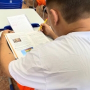 Aqui est&aacute; a descri&ccedil;&atilde;o da imagem para fins de acessibilidade 

---

**Descri&ccedil;&atilde;o da imagem "Sistema prisional do RS realiza mais de 12 mil atendimentos em educa&ccedil;&atilde;o formal e informal para pessoas privadas de liberdade.jpeg":**

Uma fotografia colorida em plano detalhe e &acirc;ngulo superior picado (visto de cima e por tr&aacute;s) foca em um detento sentado em sua carteira escolar durante uma atividade de estudo ou leitura em uma sala de aula.

**O Aluno e o Material Did&aacute;tico:**

* No centro e em primeiro plano, um homem de cabelos castanhos curt&iacute;ssimos e pele clara aparece de costas e de perfil parcial direito. Ele veste o uniforme padr&atilde;o do sistema prisional, composto por uma camiseta branca de manga curta e bermuda laranja-viva (parcialmente vis&iacute;vel na base). Ele segura um l&aacute;pis amarelo com a m&atilde;o direita, apontando para a p&aacute;gina, em uma postura concentrada de leitura. No antebra&ccedil;o esquerdo, nota-se uma pequena tatuagem minimalista escura.
* Sobre a mesa cinza com bordas azuis da carteira, h&aacute; um livro did&aacute;tico ou apostila com espiral aberto. Na p&aacute;gina esquerda do livro, h&aacute; textos impressos e duas ilustra&ccedil;&otilde;es coloridas: uma pequena imagem retangular na parte superior e, logo abaixo, a ilustra&ccedil;&atilde;o de uma bandeira com as cores do arco-&iacute;ris (s&iacute;mbolo LGBTQIA+) sob um c&eacute;u azul.
* Logo acima do livro, apoiado na aba superior da classe, h&aacute; um caderno universit&aacute;rio com espiral contendo anota&ccedil;&otilde;es manuscritas em caneta azul e vermelha.

**O Entorno e Mobili&aacute;rio:**

* &Agrave; frente e ao lado, avistam-se partes de outras carteiras escolares vazias ou ocupadas por outros detentos (um deles vis&iacute;vel ao fundo vestindo camiseta branca e outro mais distante de bermuda laranja), sugerindo uma disposi&ccedil;&atilde;o em fileiras. As cadeiras possuem encosto de pl&aacute;stico anat&ocirc;mico azul-escuro com estruturas tubulares met&aacute;licas cinzas.
* O ch&atilde;o do ambiente &eacute; revestido por lajotas cer&acirc;micas quadradas em tons de marrom e bege com linhas de rejunte escuras. A ilumina&ccedil;&atilde;o &eacute; clara, direta e uniforme.