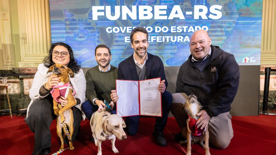 Grupo de quatro pessoas posando sobre um tapete vermelho em frente a um painel com o texto &ldquo;FUNBEA-RS&rdquo;, exibindo um documento aberto para a c&acirc;mera; tr&ecirc;s c&atilde;es aparecem em primeiro plano ao lado dos participantes.