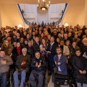 Grupo numeroso reunido no hall de um pr&eacute;dio hist&oacute;rico; v&aacute;rias pessoas ocupam o espa&ccedil;o central e os degraus, enquanto algumas utilizam cadeiras de rodas.