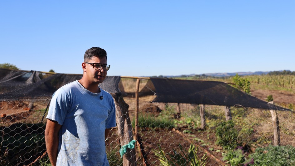 Jovem em &aacute;rea rural ao lado de uma horta cercada por tela de arame, com canteiros cultivados e estruturas cobertas por sombrite ao fundo. A paisagem inclui planta&ccedil;&otilde;es, vegeta&ccedil;&atilde;o baixa e horizonte aberto sob c&eacute;u azul sem nuvens, em um cen&aacute;rio de produ&ccedil;&atilde;o agr&iacute;cola e ilumina&ccedil;&atilde;o natural intensa.