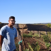 Jovem em &aacute;rea rural ao lado de uma horta cercada por tela de arame, com canteiros cultivados e estruturas cobertas por sombrite ao fundo. A paisagem inclui planta&ccedil;&otilde;es, vegeta&ccedil;&atilde;o baixa e horizonte aberto sob c&eacute;u azul sem nuvens, em um cen&aacute;rio de produ&ccedil;&atilde;o agr&iacute;cola e ilumina&ccedil;&atilde;o natural intensa.