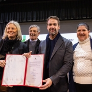 **Descri&ccedil;&atilde;o da imagem "Governo do Estado e munic&iacute;pio de Pelotas assinam conv&ecirc;nio do Programa Iconicidades para implanta&ccedil;&atilde;o de Centro de Gastronomia.jpeg":**

Uma fotografia colorida em plano m&eacute;dio e vis&atilde;o frontal mostra quatro pessoas sorrindo no palco de um audit&oacute;rio, posando para a foto enquanto exibem um documento oficial assinado.

**Os Participantes (da esquerda para a direita):**

1. Uma mulher de pele clara e cabelos loiros curtos, na altura dos ombros. Ela sorri olhando levemente para a esquerda e segura a borda esquerda de uma pasta de capa vermelha aberta. Veste um casaco sobretudo preto com um cachecol felpudo preto e cal&ccedil;a marrom.
2. Um homem mais velho de cabelos grisalhos, posicionado um pouco mais ao fundo. Ele sorri olhando na mesma dire&ccedil;&atilde;o que a primeira mulher e veste um casaco preto sobre um su&eacute;ter cinza de gola careca.
3. O governador Eduardo Leite, posicionado no centro-direito. Ele tem barba curta aparada, sorri diretamente para a c&acirc;mera e segura a borda inferior da pasta vermelha aberta. Veste um palet&oacute; cinza-escuro sobre uma blusa azul-marinho com um cachecol azul-escuro no pesco&ccedil;o.
4. Uma mulher de cabelos escuros presos em um rabo de cavalo. Ela sorri de frente para a c&acirc;mera e usa um cardig&atilde; azul-marinho aberto sobre uma blusa de tric&ocirc; branca de gola alta trabalhada com texturas, al&eacute;m de brincos dourados pendentes.

**O Documento e o Cen&aacute;rio:**

* No centro do grupo, a pasta de capa vermelha est&aacute; aberta e voltada para a frente, revelando em sua p&aacute;gina direita um documento oficial impresso em papel branco com o bras&atilde;o do Estado no topo e assinaturas com carimbos na parte inferior. A p&aacute;gina esquerda da pasta est&aacute; em branco.
* Ao fundo, uma tela de proje&ccedil;&atilde;o clara exibe parcialmente a palavra **"V&Ecirc;NIO"** em letras escuras e mai&uacute;sculas.
* Na parte superior e nas laterais do palco, h&aacute; cortinas e franjas de tecido cinza-escuro dispostas verticalmente. No canto extremo esquerdo, avista-se um detalhe de um totem branco com o logotipo oficial do **"GOVERNO DO ESTADO RIO GRANDE DO SUL"**. A ilumina&ccedil;&atilde;o &eacute; frontal e focada no grupo.