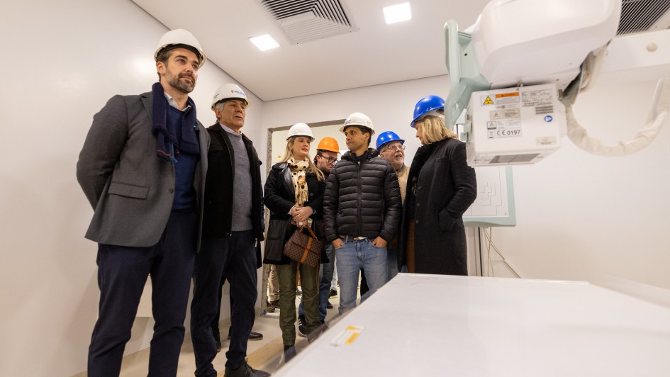 Sala hospitalar em fase de instala&ccedil;&atilde;o, com equipamento de imagem m&eacute;dica em primeiro plano. Um grupo de visitantes usando capacetes observa o ambiente, que possui paredes claras, ilumina&ccedil;&atilde;o forte e acabamento novo.