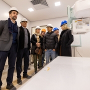 Sala hospitalar em fase de instala&ccedil;&atilde;o, com equipamento de imagem m&eacute;dica em primeiro plano. Um grupo de visitantes usando capacetes observa o ambiente, que possui paredes claras, ilumina&ccedil;&atilde;o forte e acabamento novo.