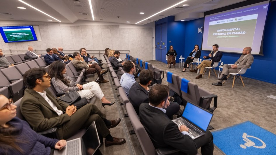 Descri&ccedil;&atilde;o da imagem "Road show na B3 abriu fase de apresenta&ccedil;&atilde;o ao mercado para estudo do projeto que viabiliza institui&ccedil;&atilde;o com 350 leitos 100% SUS.jpeg":

Uma fotografia colorida em plano geral e &acirc;ngulo levemente picado mostra o interior de um audit&oacute;rio moderno durante uma apresenta&ccedil;&atilde;o institucional. A plateia est&aacute; sentada em poltronas dispostas em fileiras inclinadas &agrave; esquerda, enquanto os palestrantes ocupam uma &aacute;rea de palco &agrave; direita.

O Palco e os Palestrantes (&agrave; direita):
&Agrave; frente de uma parede azul-escura com o logotipo branco da "[B]&sup3;" (Bolsa de Valores do Brasil), quatro pessoas est&atilde;o sentadas em cadeiras brancas de estilo moderno com pernas de madeira clara, dispostas em um semic&iacute;rculo:

Da esquerda para a direita: Uma mulher com blusa escura e saia marrom; uma mulher de terno escuro e camisa social clara; o governador Eduardo Leite, vestindo um palet&oacute; escuro sobre camisa branca, cal&ccedil;a bege e sapatos marrons, segurando um microfone e gesticulando enquanto fala; e um homem calvo vestindo um terno cinza ou bege-claro, olhando em dire&ccedil;&atilde;o ao governador.

Atr&aacute;s deles, uma grande tela de proje&ccedil;&atilde;o exibe um slide com o t&iacute;tulo principal: "NOVO HOSPITAL ESTADUAL EM VIAM&Atilde;O - APRESENTA&Ccedil;&Atilde;O DO PROJETO". O slide possui logotipos institucionais no topo, incluindo o do BID (Banco Interamericano de Desenvolvimento) e o do Governo do Estado do Rio Grande do Sul.

A Plateia (&agrave; esquerda e ao centro):
V&aacute;rias pessoas em trajes formais e casuais de neg&oacute;cios assistem &agrave; apresenta&ccedil;&atilde;o.

Em primeiro plano, no canto inferior esquerdo, uma mulher usa &oacute;culos e digita em um notebook prata. Ao lado dela, um homem de &oacute;culos e palet&oacute; verde-oliva acompanha a fala. Atr&aacute;s deles, uma mulher com blusa estampada em preto e branco e cal&ccedil;a branca apoia uma bolsa preta no colo.

Nas fileiras centrais, um homem de terno escuro, sentado de costas para a c&acirc;mera, trabalha em um notebook com a tela azul ligada.

As poltronas do audit&oacute;rio s&atilde;o estofadas em tom cinza-escuro. Algumas poltronas vazias nas fileiras centrais possuem capas azuis nos encostos. No canto superior esquerdo, h&aacute; uma segunda tela menor fixada na parede exibindo o mesmo slide da apresenta&ccedil;&atilde;o.

O Ambiente:
O audit&oacute;rio possui piso acarpetado cinza. No canto inferior direito, h&aacute; um tapete azul retangular com o s&iacute;mbolo internacional de acessibilidade para cadeirantes em branco. O teto &eacute; composto por pain&eacute;is texturizados claros com linhas embutidas de ilumina&ccedil;&atilde;o LED e lumin&aacute;rias spot acesas, criando uma atmosfera clara e profissional.