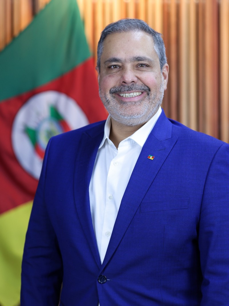 Foto em plano americano de um homem de meia idade, de cabelo e barba grisalhos. De terno azul, ele sorri. Ao fundo, aparece parte da bandeira do RS, nas cores verde, vermelha e amarela.