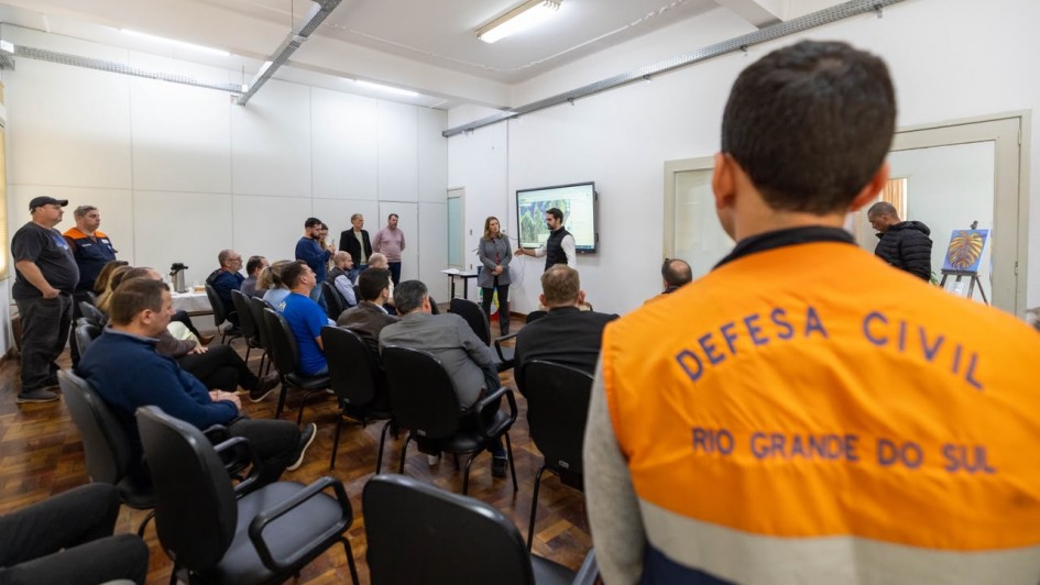 V&aacute;rias pessoas est&atilde;o emuma grande sala. Em primeiro pano, um home de costas vestindo um jaleco com os dizeres Defesa Civil Rio Grande do Sul.