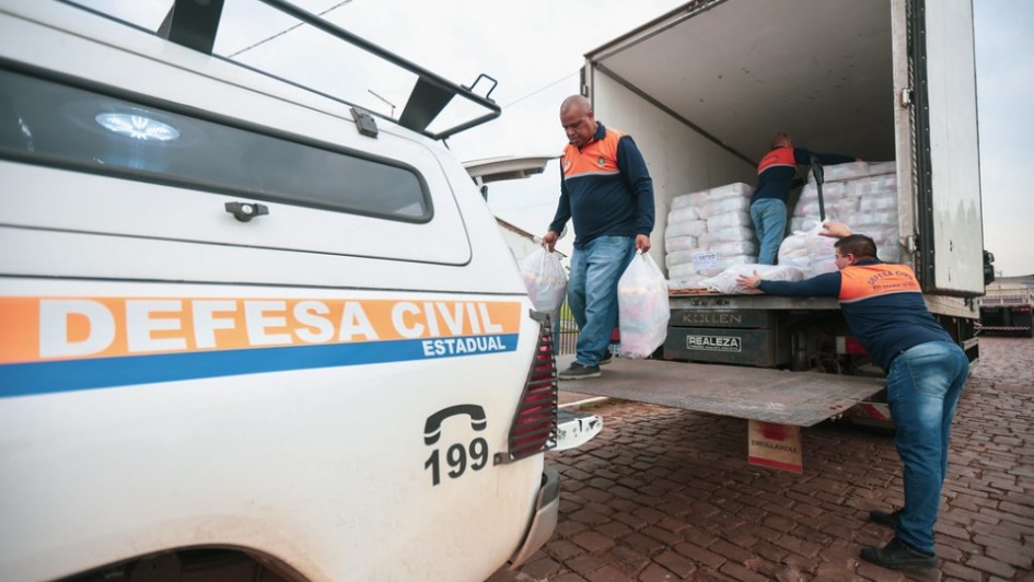

Uma fotografia colorida em plano m&eacute;dio e perspectiva em contra-plong&eacute;e (de baixo para cima) mostra uma opera&ccedil;&atilde;o de descarregamento de doa&ccedil;&otilde;es ou suprimentos de ajuda humanit&aacute;ria realizada por agentes da Defesa Civil.

**Os Agentes e a A&ccedil;&atilde;o:**

* Tr&ecirc;s homens vestindo o uniforme operacional da Defesa Civil &mdash; camisetas de mangas compridas em azul-marinho com ombros e gola em laranja, e cal&ccedil;as jeans &mdash; participam do trabalho.
* No centro, um homem caminha sobre a plataforma elevat&oacute;ria traseira do caminh&atilde;o, carregando dois grandes sacos pl&aacute;sticos transparentes cheios de suprimentos, um em cada m&atilde;o.
* &Agrave; direita, em primeiro plano, outro agente est&aacute; em p&eacute; na rua, esticando os bra&ccedil;os para receber um pacote do interior do ve&iacute;culo.
* Mais ao fundo, no interior do ba&uacute; do caminh&atilde;o, um terceiro agente organiza fardos empilhados de doa&ccedil;&otilde;es mantidas em embalagens pl&aacute;sticas transparentes.

**Os Ve&iacute;culos e o Ambiente:**

* No canto esquerdo, em primeiro plano, destaca-se a lateral traseira de uma caminhonete branca da Defesa Civil. Na lataria, h&aacute; uma faixa horizontal laranja e azul com a inscri&ccedil;&atilde;o em letras mai&uacute;sculas **"DEFESA CIVIL ESTADUAL"** e o n&uacute;mero de emerg&ecirc;ncia **"199"** acompanhado do &iacute;cone de um telefone em preto. No teto do ve&iacute;culo, v&ecirc;-se um bagageiro de metal preto.
* &Agrave; direita, o caminh&atilde;o-ba&uacute; de carga est&aacute; com as portas traseiras totalmente abertas e a plataforma hidr&aacute;ulica abaixada ao n&iacute;vel de acesso.
* O solo &eacute; de pavimento de paralelep&iacute;pedos em tons avermelhados e o c&eacute;u ao fundo est&aacute; nublado e claro.