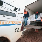

Uma fotografia colorida em plano m&eacute;dio e perspectiva em contra-plong&eacute;e (de baixo para cima) mostra uma opera&ccedil;&atilde;o de descarregamento de doa&ccedil;&otilde;es ou suprimentos de ajuda humanit&aacute;ria realizada por agentes da Defesa Civil.

**Os Agentes e a A&ccedil;&atilde;o:**

* Tr&ecirc;s homens vestindo o uniforme operacional da Defesa Civil &mdash; camisetas de mangas compridas em azul-marinho com ombros e gola em laranja, e cal&ccedil;as jeans &mdash; participam do trabalho.
* No centro, um homem caminha sobre a plataforma elevat&oacute;ria traseira do caminh&atilde;o, carregando dois grandes sacos pl&aacute;sticos transparentes cheios de suprimentos, um em cada m&atilde;o.
* &Agrave; direita, em primeiro plano, outro agente est&aacute; em p&eacute; na rua, esticando os bra&ccedil;os para receber um pacote do interior do ve&iacute;culo.
* Mais ao fundo, no interior do ba&uacute; do caminh&atilde;o, um terceiro agente organiza fardos empilhados de doa&ccedil;&otilde;es mantidas em embalagens pl&aacute;sticas transparentes.

**Os Ve&iacute;culos e o Ambiente:**

* No canto esquerdo, em primeiro plano, destaca-se a lateral traseira de uma caminhonete branca da Defesa Civil. Na lataria, h&aacute; uma faixa horizontal laranja e azul com a inscri&ccedil;&atilde;o em letras mai&uacute;sculas **"DEFESA CIVIL ESTADUAL"** e o n&uacute;mero de emerg&ecirc;ncia **"199"** acompanhado do &iacute;cone de um telefone em preto. No teto do ve&iacute;culo, v&ecirc;-se um bagageiro de metal preto.
* &Agrave; direita, o caminh&atilde;o-ba&uacute; de carga est&aacute; com as portas traseiras totalmente abertas e a plataforma hidr&aacute;ulica abaixada ao n&iacute;vel de acesso.
* O solo &eacute; de pavimento de paralelep&iacute;pedos em tons avermelhados e o c&eacute;u ao fundo est&aacute; nublado e claro.
