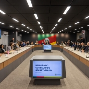Uma fotografia colorida em plano geral e perspectiva central sim&eacute;trica mostra uma sala de reuni&otilde;es ampla e moderna durante um encontro de trabalho presencial.

**A Sala e a Disposi&ccedil;&atilde;o das Mesas:**

* As mesas de tom madeira clara com acabamento cinza-escuro est&atilde;o dispostas em formato de ret&acirc;ngulo aberto (em U).
* Nas duas laterais compridas e ao fundo da mesa, dezenas de secret&aacute;rios e assessores est&atilde;o sentados em cadeiras escuras, acompanhando a reuni&atilde;o. Diversos participantes utilizam notebooks, blocos de notas ou copos/garrafas de &aacute;gua sobre a mesa.
* No centro da cabeceira (ao fundo), encontra-se a mesa de honra, encabe&ccedil;ada pelo governador Eduardo Leite e pelo vice-governador Gabriel Souza, acompanhados por outros secret&aacute;rios e autoridades em trajes formais e coletes operacionais.
* Ao fundo da sala, centralizada na parede cinza-escura, destaca-se uma grande imagem da bandeira do Estado do Rio Grande do Sul (nas cores verde, vermelha e amarela, com o bras&atilde;o estadual ao centro).

**Monitores e Elementos Visuais:**

* Em primeiro plano, no centro da imagem, h&aacute; um monitor retangular posicionado em um pedestal cinza voltado para a entrada da sala. A tela exibe o t&iacute;tulo em letras azuis mai&uacute;sculas: **"REUNI&Atilde;O DO CONSELHO DE SECRETARIADO"**, com a data e o logotipo do Governo do Estado do Rio Grande do Sul na parte inferior.
* Um segundo monitor com a mesma apresenta&ccedil;&atilde;o gr&aacute;fica est&aacute; centralizado no interior do formato em U das mesas.
* Nas paredes laterais da sala, telas adicionais fixadas na parede reproduzem o mesmo slide da apresenta&ccedil;&atilde;o.

**Infraestrutura e Ilumina&ccedil;&atilde;o:**

* O teto &eacute; preto e conta com diversas calhas de ilumina&ccedil;&atilde;o LED cont&iacute;nuas dispostas em linhas paralelas na vertical, conferindo alta luminosidade ao ambiente.
* O piso &eacute; coberto por um carpete cinza-claro com textura discreta. No canto direito da sala, ao fundo, observa-se um profissional de imprensa em p&eacute; com uma c&acirc;mera fotogr&aacute;fica apontada para a reuni&atilde;o.