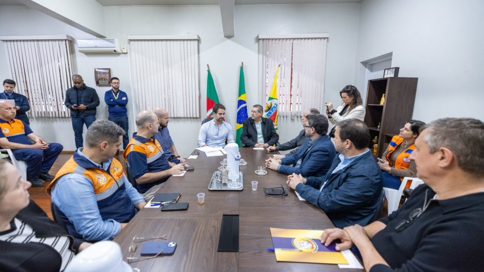 **Descri&ccedil;&atilde;o da imagem "Reuni&otilde;es ocorreram nesta quinta (30) Cr&eacute;dito Vitor RosaSecom.jpeg":**

Uma fotografia colorida em plano m&eacute;dio e perspectiva angular mostra uma reuni&atilde;o de trabalho em torno de uma mesa retangular de madeira escura.

**Integrantes e Disposi&ccedil;&atilde;o na Mesa:**

* Ao fundo, no centro da mesa, o governador Eduardo Leite veste camisa social azul-clara e fala aos presentes com as m&atilde;os espalmadas sobre pap&eacute;is.
* &Agrave; sua esquerda (&agrave; direita no enquadramento), est&aacute; um homem de terno escuro e camisa cinza.
* Ao longo das duas laterais da mesa retangular, cerca de dez pessoas &mdash; entre homens e mulheres &mdash; acompanham a reuni&atilde;o.
* Do lado esquerdo da mesa, alguns participantes vestem coletes operacionais e uniformes da Defesa Civil (nas cores laranja e azul-marinho com cinza).
* Do lado direito e em primeiro plano, homens e mulheres usam trajes casuais e formais de inverno, como palet&oacute;s, jaquet&otilde;es e blusas pretas.
* Sobre a mesa, h&aacute; uma garrafa t&eacute;rmica branca com copos descart&aacute;veis em uma bandeja de inox, al&eacute;m de cadernos de anota&ccedil;&otilde;es, &oacute;culos de grau, telefones celulares e uma pasta amarela com o bras&atilde;o do Estado.

**O Ambiente e Elementos ao Fundo:**

* Diretamente atr&aacute;s do governador, est&atilde;o hasteadas tr&ecirc;s bandeiras em mastros de madeira: a bandeira do Estado do Rio Grande do Sul (esquerda), a bandeira do Brasil (centro) e a bandeira de um munic&iacute;pio ga&uacute;cho (direita).
* A sala possui paredes em tom cinza-claro, vigas no teto e janelas ao fundo cobertas por persianas verticais beges.
* Ao fundo, do lado esquerdo, tr&ecirc;s homens est&atilde;o em p&eacute; observando a reuni&atilde;o. Do lado direito, uma mulher segura um smartphone para registrar o evento e outra pessoa de colete da Defesa Civil assiste &agrave; reuni&atilde;o sentada em uma cadeira branca.