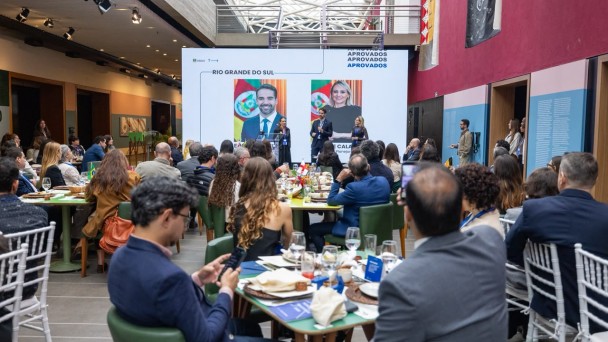 A imagem mostra um sal&atilde;o com mesas redondas, com diversas pessoas sentadas olhando para o palco, onde se encontra o governador falando ao microfone e outras duas mulheres. Ao fundo, h&aacute; um painel iluminado com fotos do governador e da titular da SPGG e as palavras "Aprovados" e "Rio Grande do Sul". 