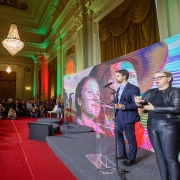 Foto de sal&atilde;o de estilo cl&aacute;ssico, com tapete vermelho. Um homem fala ao p&uacute;lpito no canto direito, em trajes formais. A plateia est&aacute; cheia e ocupa o lado esquerdo e o fundo do quadro, com a maior parte das pessoas sentada.