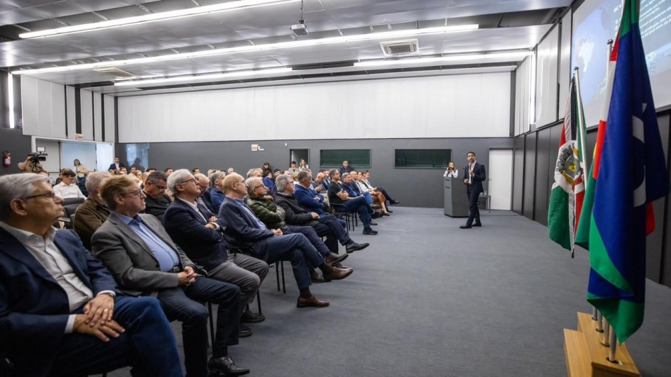 Foto de um evento em ambiente fechado e formal, de paredes brancas e piso cinza. &Aacute; direita, Leite aparece de p&eacute; falando ao microfone. &Agrave; esquerda, a plateia acompanha a apresenta&ccedil;&atilde;o, sentada.