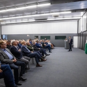 Foto de um evento em ambiente fechado e formal, de paredes brancas e piso cinza. &Aacute; direita, Leite aparece de p&eacute; falando ao microfone. &Agrave; esquerda, a plateia acompanha a apresenta&ccedil;&atilde;o, sentada.