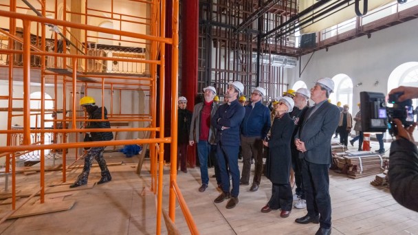 Uma fotografia colorida em plano geral e &acirc;ngulo levemente contra-plong&eacute;e (de baixo para cima) mostra uma comitiva de autoridades e t&eacute;cnicos em visita &agrave;s obras de restaura&ccedil;&atilde;o no interior do Theatro S&atilde;o Pedro.

As Pessoas e a Visita:

Ao centro e &agrave; direita, um grupo de homens e mulheres usa capacetes de prote&ccedil;&atilde;o brancos para obras e trajes sociais e casuais de inverno. Todos olham para o alto e para a frente, observando os detalhes da estrutura do teto e do palco.

Entre eles, destacam-se:

O governador Eduardo Leite, posicionado ao centro, vestindo palet&oacute; azul-marinho sobre camisa clara, cal&ccedil;a escura e sapatos marrons, com as m&atilde;os cruzadas &agrave; frente.

Ao lado dele, um homem mais velho com casaco cinza aberto e blusa de gola alta vermelha.

&Agrave; direita, uma mulher de sobretudo preto e &oacute;culos, um homem de camiseta preta estampada, e outro homem de palet&oacute; cinza-escuro com camisa escura e sapatos pretos.

&Agrave; esquerda, em meio &agrave;s estruturas met&aacute;licas, uma mulher usando capacete amarelo e cal&ccedil;a camuflada caminha pelo piso da obra.

No canto inferior direito, v&ecirc;-se em primeiro plano a m&atilde;o e a c&acirc;mera de v&iacute;deo de um cinegrafista registrando o evento.

O Ambiente e as Obras:

O grupo est&aacute; sobre o tablado de t&aacute;buas de madeira crua do palco.

Do lado esquerdo, ergue-se uma alta torre de andaimes met&aacute;licos tubulares pintados de laranja-vivo, que alcan&ccedil;a os camarotes e as galerias cl&aacute;ssicas do teatro ao fundo.

Ao centro, h&aacute; uma grande cortina de veludo vermelho recolhida.

Acima do grupo, observa-se toda a estrutura c&ecirc;nica superior exposta, com vigas de a&ccedil;o pretas, varas de ilumina&ccedil;&atilde;o, urupemas e cabos de sustenta&ccedil;&atilde;o.

&Agrave; direita, a parede branca do teatro possui grandes janelas em arco com molduras brancas e, no ch&atilde;o, pilhas de ripas de madeira e t&aacute;buas prontas para instala&ccedil;&atilde;o.