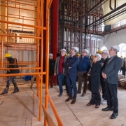 Uma fotografia colorida em plano geral e &acirc;ngulo levemente contra-plong&eacute;e (de baixo para cima) mostra uma comitiva de autoridades e t&eacute;cnicos em visita &agrave;s obras de restaura&ccedil;&atilde;o no interior do Theatro S&atilde;o Pedro.

As Pessoas e a Visita:

Ao centro e &agrave; direita, um grupo de homens e mulheres usa capacetes de prote&ccedil;&atilde;o brancos para obras e trajes sociais e casuais de inverno. Todos olham para o alto e para a frente, observando os detalhes da estrutura do teto e do palco.

Entre eles, destacam-se:

O governador Eduardo Leite, posicionado ao centro, vestindo palet&oacute; azul-marinho sobre camisa clara, cal&ccedil;a escura e sapatos marrons, com as m&atilde;os cruzadas &agrave; frente.

Ao lado dele, um homem mais velho com casaco cinza aberto e blusa de gola alta vermelha.

&Agrave; direita, uma mulher de sobretudo preto e &oacute;culos, um homem de camiseta preta estampada, e outro homem de palet&oacute; cinza-escuro com camisa escura e sapatos pretos.

&Agrave; esquerda, em meio &agrave;s estruturas met&aacute;licas, uma mulher usando capacete amarelo e cal&ccedil;a camuflada caminha pelo piso da obra.

No canto inferior direito, v&ecirc;-se em primeiro plano a m&atilde;o e a c&acirc;mera de v&iacute;deo de um cinegrafista registrando o evento.

O Ambiente e as Obras:

O grupo est&aacute; sobre o tablado de t&aacute;buas de madeira crua do palco.

Do lado esquerdo, ergue-se uma alta torre de andaimes met&aacute;licos tubulares pintados de laranja-vivo, que alcan&ccedil;a os camarotes e as galerias cl&aacute;ssicas do teatro ao fundo.

Ao centro, h&aacute; uma grande cortina de veludo vermelho recolhida.

Acima do grupo, observa-se toda a estrutura c&ecirc;nica superior exposta, com vigas de a&ccedil;o pretas, varas de ilumina&ccedil;&atilde;o, urupemas e cabos de sustenta&ccedil;&atilde;o.

&Agrave; direita, a parede branca do teatro possui grandes janelas em arco com molduras brancas e, no ch&atilde;o, pilhas de ripas de madeira e t&aacute;buas prontas para instala&ccedil;&atilde;o.