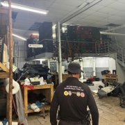 Uma fotografia colorida em plano geral e &acirc;ngulo normal mostra o interior de um grande galp&atilde;o ou ferro-velho de autope&ccedil;as durante uma opera&ccedil;&atilde;o de fiscaliza&ccedil;&atilde;o.

O Agente em Primeiro Plano:

No centro da imagem, em primeiro plano e de costas para a c&acirc;mera, destaca-se um agente policial ou de fiscaliza&ccedil;&atilde;o.

Ele veste um uniforme t&aacute;tico composto por blusa preta de manga longa com a inscri&ccedil;&atilde;o em letras mai&uacute;sculas brancas nas costas: "SECRETARIA DA SEGURAN&Ccedil;A P&Uacute;BLICA".

Abaixo do texto, h&aacute; uma fileira de quatro bras&otilde;es e distintivos coloridos institucionais (incluindo os s&iacute;mbolos do Estado do RS, Pol&iacute;cia Civil, DetranRS e Instituto-Geral de Per&iacute;cias).

Ele usa um chap&eacute;u t&aacute;tico do tipo bucket/saf&aacute;ri preto e cinto com coldre e equipamentos operacionais na cintura.

O Interior do Estabelecimento:

O espa&ccedil;o &eacute; amplo e possui p&eacute;-direito alto, piso de concreto/cer&acirc;mica desgastada e paredes de blocos com divis&oacute;rias de chapas met&aacute;licas.

&Agrave; esquerda, h&aacute; estantes de madeira r&uacute;stica e bancadas abarrotadas de pe&ccedil;as de ve&iacute;culos, como portas de carros, pain&eacute;is, blocos de motor, estofamentos de bancos e rolos de pl&aacute;stico-bolha. Uma placa branca suspensa traz a indica&ccedil;&atilde;o "Montagem" com uma seta apontada para a esquerda.

Ao fundo e no mezanino met&aacute;lico superior, h&aacute; centenas de caixas pl&aacute;sticas organizadoras (pretas, verdes e vermelhas) empilhadas at&eacute; o teto, al&eacute;m de ferragens e eixos veiculares suspensos.

&Agrave; direita, uma escada met&aacute;lica aberta d&aacute; acesso ao mezanino superior. No piso t&eacute;rreo ao lado da escada, outro agente ou operador agacha-se pr&oacute;ximo a bancos automotivos e blocos de motor espalhados pelo ch&atilde;o para inspecionar as pe&ccedil;as. A ilumina&ccedil;&atilde;o &eacute; fornecida por lumin&aacute;rias tubulares fluorescentes e l&acirc;mpadas de LED no teto de forro modular.
