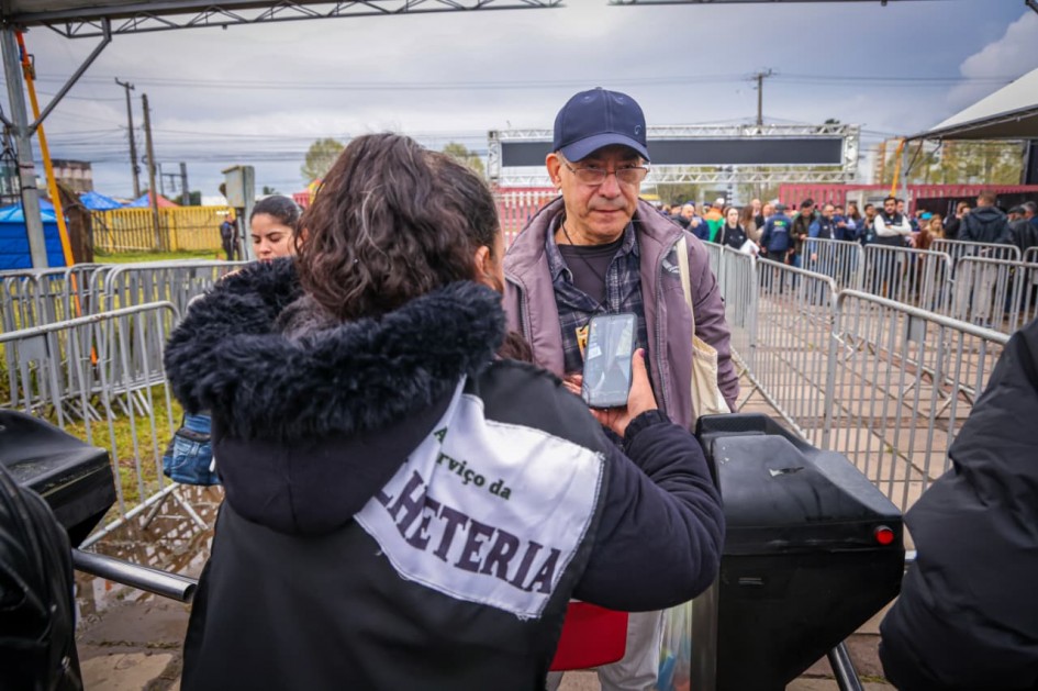 Uma fotografia colorida em primeiro plano e ângulo frontal registra a entrada de um visitante passando pela catraca de controle de acesso de um evento ao ar livre.<br />
<br />
O Visitante e a Atendente:<br />
<br />
No centro da imagem, um homem de pele clara e meia-idade veste um boné de aba curva azul-marinho, óculos de grau retangulares, jaqueta corta-vento em tom roxo-acinzentado/beringela sobre camisa xadrez escura e uma bolsa de tecido clara a tiracolo. Ele olha para baixo com expressão tranquila enquanto aguarda a liberação.<br />
<br />
Em primeiro plano, de costas para a câmera, uma atendente com cabelos escuros presos e mechas grisalhas usa um casaco preto com capuz peludo. Sobre o casaco, há um tecido ou colete branco com a inscrição em letras maiúsculas: "A serviço da BILHETERIA". Ela segura um smartphone verticalmente para escanear ou conferir o ingresso digital apresentado pelo visitante.<br />
<br />
A Catraca e o Acesso:<br />
<br />
Entre a atendente e o homem, vê-se a estrutura preta de uma catraca eletrônica com braços metálicos giratórios e uma luz indicadora vermelha na lateral.<br />
<br />
Ao lado da atendente (à esquerda), o rosto de outra mulher de cabelos escuros aparece parcialmente no enquadramento.<br />
<br />
O Entorno e o Fundo:<br />
<br />
A área de acesso é cercada por gradis e cercas de contenção metálicas cinzas dispostas em corredores organizadores de fluxo.<br />
<br />
Ao fundo, vê-se uma multidão de pessoas em fila aguardando para entrar, sob uma grande estrutura de pórtico metálico e tendas brancas.<br />
<br />
O céu está completamente encoberto por nuvens cinzentas, indicando tempo frio e nublado. No solo de paralelepípedos e lajotas, observam-se poças dágua e lama próximas à grama molhada.