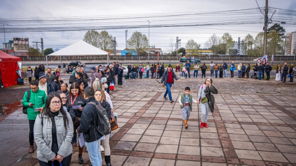 Uma fotografia colorida em plano geral e ângulo levemente picado captura um grande grupo de pessoas reunidas em uma praça pública pavimentada com lajotas de pedra em tons de bege e marrom.<br />
<br />
**Primeiro Plano:**<br />
<br />
* No canto inferior esquerdo, uma fila de pessoas se forma em direção ao fundo da imagem. Em destaque na frente, uma mulher de pele clara e óculos de armação preta veste um casaco cinza-claro estruturado sobre camisa preta, segurando uma bolsa azul na mão esquerda. Atrás dela, uma mulher de cabelos pretos e jaqueta de couro preta olha para a câmera. Mais atrás na fila, destaca-se um homem com jaqueta esportiva verde-limão da Adidas.<br />
* No centro-direito, um menino pequeno em trajes tradicionais gaúchos (bombacha cinza, botas de couro marrom, camisa clara e colete verde com lenço vermelho no pescoço) caminha de mãos dadas com uma mulher de moletom cinza e óculos, que fala ao celular com a mão direita. À frente deles, um homem de camiseta vermelha e calça jeans caminha no sentido oposto.<br />
<br />
**Meio Plano e Fundo:**<br />
<br />
* Uma extensa fileira de dezenas de pessoas estende-se da esquerda para a direita, acompanhando a borda de uma rua ou acostamento ao fundo. Elas estão em pé, de costas ou perfil, aguardando algo.<br />
* Do lado esquerdo, vê-se uma grande barraca ou tenda de lona vermelha e, logo ao lado, uma tenda branca pontiaguda maior, com algumas pessoas próximas e uma caminhonete branca estacionada sob a cobertura.<br />
* A paisagem de fundo inclui uma rua asfáltica, canteiros de vegetação rasteira e algumas árvores com copas em tons de verde-claro e amarelo. À direita, alinham-se mastros de bandeiras brancas e azuis com logotipos institucionais.<br />
* O céu está completamente encoberto por nuvens cinzentas e densas. O piso de lajotas está ligeiramente molhado e irregular.