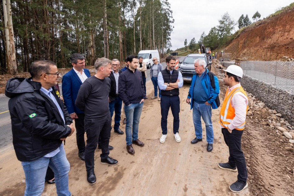 Uma fotografia colorida em plano geral e ângulo levemente picado registra uma vistoria técnica às margens de uma rodovia em obras de contenção de encosta.
**A Comitiva e os Técnicos (em primeiro e meio plano):**
* No centro da composição, um grupo de autoridades e engenheiros forma uma roda de conversa sobre o acostamento de terra batida da rodovia.
* Ao centro, destaca-se o governador Eduardo Leite, de braços cruzados, vestindo colete acolchoado preto sobre camisa social clara de mangas compridas dobradas, calça escura e tênis brancos.
* À direita, em primeiro plano de perfil, um engenheiro ou fiscal de obra usa capacete de proteção branco, camisa clara com colete refletivo laranja de faixas cinzas, calça preta e tênis escuros. Ao lado dele, um homem mais velho de cabelos brancos gesticula enquanto fala, vestindo jaqueta esportiva azul-royal sobre suéter azul, calça jeans e sapatos pretos.
* À esquerda, completam o grupo gestores e assessores: um homem de costas parciais em jaqueta preta com a bandeira do Brasil no braço esquerdo, outro com suéter cinza-escuro sobre camiseta, e autoridades em paletó azul-marinho e jaquetas escuras.
**O Local da Obra e o Entorno:**
* À direita, ao lado do acostamento, ergue-se uma barreira de contenção recém-construída em gabião (estruturas em gaiolas de tela de arame preenchidas com pedras brutas), sustentando uma encosta íngreme de terra vermelha exposta e cortada pela obra.
* Ao fundo da pista, avista-se uma caminhonete preta estacionada, uma van branca e operários trabalhando próximos a uma retroescavadeira na margem da estrada.
* À esquerda da pista simples de asfalto, estende-se uma floresta densa e vertical de eucaliptos altos sob um céu encoberto por nuvens cinzentas claras.