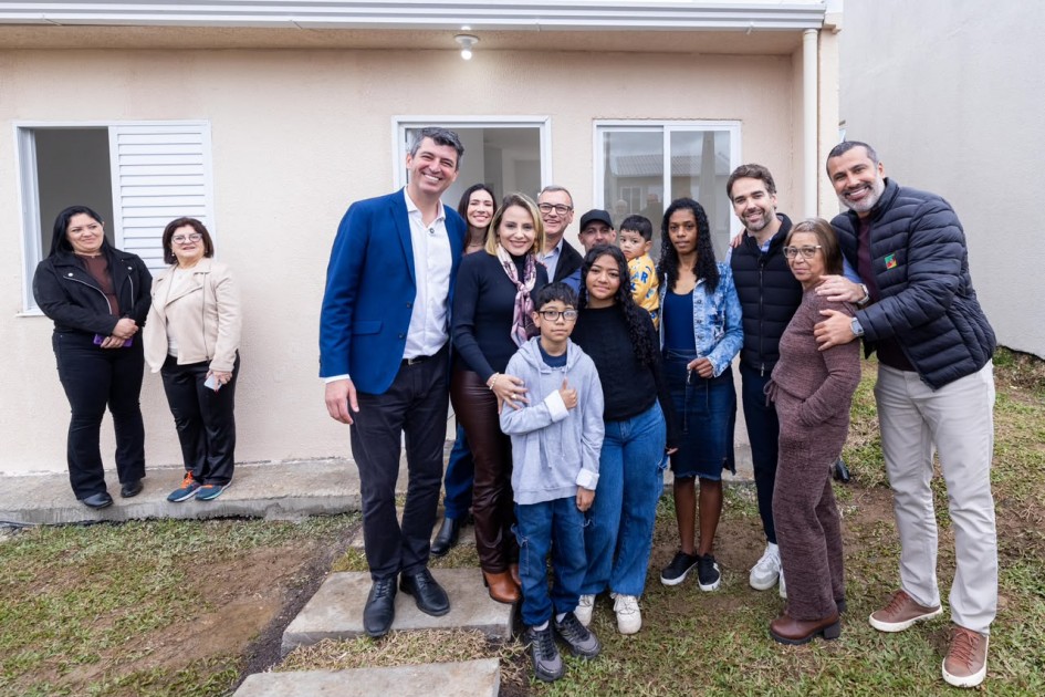 Uma fotografia colorida em plano inteiro e ângulo frontal retrata a entrega de uma casa popular, com autoridades estaduais posando para foto ao lado da família beneficiada em frente ao imóvel recém-construído.
**As Pessoas (ao centro e em primeiro plano):**
* Ao centro, a família e os gestores reúnem-se sorrindo em um momento comemorativo.
* À esquerda do grupo central, um homem alto de blazer azul-marinho, camisa branca e calça preta sorri amplamente. Ao lado dele, uma mulher de blusa preta, lenço estampado no pescoço e calça marrom abraça um menino de óculos e moletom cinza com capuz.
* Ao lado do garoto, uma jovem de blusa preta e calça jeans sorri para a foto, próxima a uma mulher de jaqueta jeans com saia azul segurando uma criança pequena nos braços.
* À direita, o governador Eduardo Leite, vestindo colete acolchoado preto sobre camisa social clara e calça escura, sorri ao lado de uma senhora de conjunto marrom.
* No extremo direito, um homem de pele morena, barba e cabelo curto raspado veste jaqueta acolchoada preta com pequeno emblema da bandeira do Rio Grande do Sul no peito, calça bege e tênis marrom, abraçando a senhora pelos ombros.
* À esquerda, em segundo plano, duas mulheres acompanham a cerimônia próximas à janela: uma de jaqueta de couro preta e calça preta, e outra de blazer bege-claro e calça escura.
**A Casa e o Entorno:**
* O grupo está posicionado sobre uma calçada de blocos de concreto e gramado rasteiro recém-plantado em frente à residência térrea.
* A casa possui paredes pintadas em tom bege-claro/areia, com porta de entrada de madeira, janela de correr de vidro transparente com esquadria de alumínio branco e outra janela com veneziana branca aberta à esquerda. Uma lâmpada simples acesa ilumina a entrada sob a marquise.