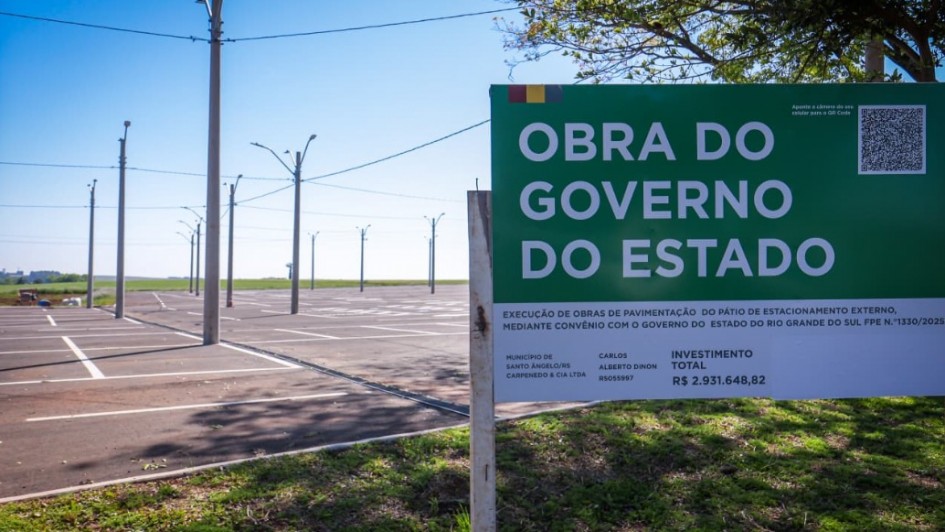 Uma fotografia colorida em plano médio e ângulo frontal foca em uma placa oficial de obra pública instalada ao lado de um amplo pátio de estacionamento recém-pavimentado.

**A Placa de Obra (em primeiro plano à direita):**

* No lado direito da imagem, sobressai uma grande placa retangular com topo em tom verde-escuro e base branca.
* No topo verde, lê-se em letras garrafais brancas maiúsculas: **"OBRA DO GOVERNO DO ESTADO"**, com uma pequena bandeira do Rio Grande do Sul no canto superior esquerdo e um QR Code no canto superior direito.
* Na parte inferior branca, constam os detalhes técnicos e institucionais da intervenção:
* **Descrição:** *"EXECUÇÃO DE OBRAS DE PAVIMENTAÇÃO DO PÁTIO DE ESTACIONAMENTO EXTERNO, MEDIANTE CONVÊNIO COM O GOVERNO DO ESTADO DO RIO GRANDE DO SUL FPE N.º 1330/2025"*.
* **Município:** *"SANTO ÂNGELO/RS"*.
* **Empresa Executora:** *"CARPENEDO & CIA LTDA"*.
* **Investimento Total:** *"R$ 2.931.648,82"*.



**O Estacionamento e o Entorno (à esquerda e ao fundo):**

* À esquerda, estende-se o amplo pátio de asfalto novo e escuro, marcado por demarcações brancas de vagas de estacionamento recém-pintadas.
* Ao longo do pátio, alinham-se altos postes de concreto cinza com luminárias de iluminação pública e fiação disposta no topo.
* Ao fundo, avista-se uma vegetação rasteira e terreno plano sob um céu limpo de tom azul-vivo, sob iluminação solar direta. 
