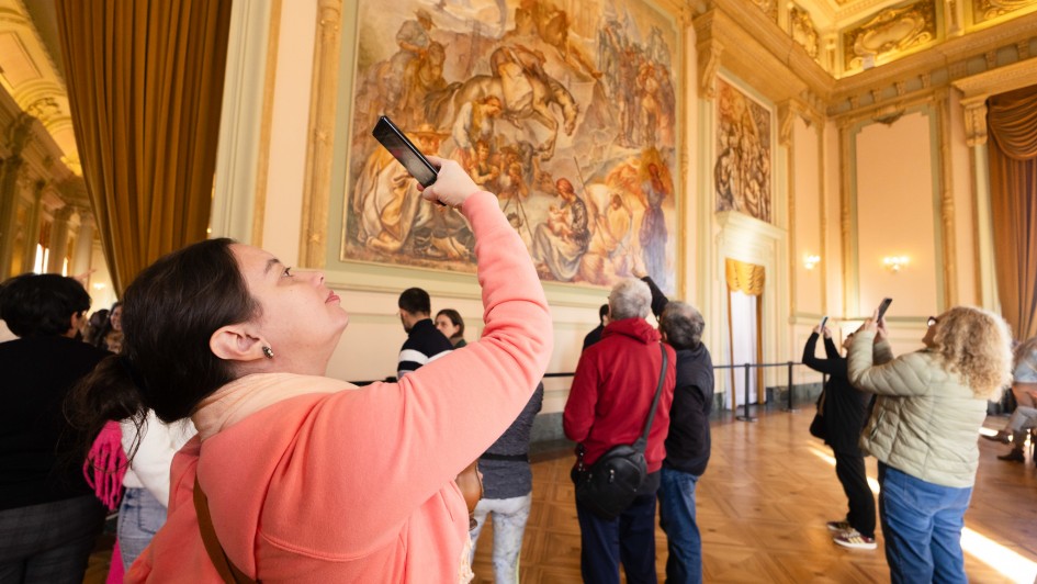 Uma fotografia colorida em plano m&eacute;dio e &acirc;ngulo contraponto (de baixo para cima) regista visitantes a apreciar e fotografar obras de arte no interior de um sal&atilde;o cl&aacute;ssico do Pal&aacute;cio Piratini.

**Em Primeiro Plano (&agrave; esquerda):**

* Em destaque em primeiro plano e de perfil, uma mulher de pele clara e cabelo escuro apanhado num coque ergue o seu smartphone com a m&atilde;o direita para fotografar o teto e as pinturas. Ela veste um casaco com capuz em tom rosa-salm&atilde;o/coral e traz uma mala a tiracolo com al&ccedil;a castanha.



**Os Visitantes e as Obras de Arte (ao centro e &agrave; direita):**

* Ao centro e &agrave; direita, outros visitantes observam e fotografam os afrescos nas paredes e no teto:


* Um homem de casaco vermelho e bolsa tiracolo preta est&aacute; de costas a admirar um grande painel pintado.


* Ao lado dele, um homem de casaco escuro aponta na dire&ccedil;&atilde;o da pintura.


* Mais &agrave; direita, duas mulheres erguem os seus telem&oacute;veis para capturar imagens do ambiente. Uma delas veste um casaco acolchoado bege e cal&ccedil;as jeans azuis.





**A Arquitetura do Sal&atilde;o (ao fundo):**

* A parede oposta exibe um imponente painel pintado cl&aacute;ssico em tons de bege, terra e azul, retratando figuras hist&oacute;ricas e cavalos.


* O sal&atilde;o nobre apresenta arquitetura cl&aacute;ssica rica em detalhes: teto trabalhado com molduras douradas e afrescos, colunas e pilastras ornamentadas, ilumina&ccedil;&atilde;o quente e piso de parquet de madeira polida.


* &Agrave; esquerda, sobressaem-se volumosas cortinas de veludo em tom dourado.