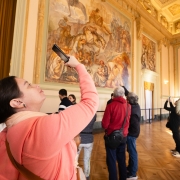 Uma fotografia colorida em plano m&eacute;dio e &acirc;ngulo contraponto (de baixo para cima) regista visitantes a apreciar e fotografar obras de arte no interior de um sal&atilde;o cl&aacute;ssico do Pal&aacute;cio Piratini.

**Em Primeiro Plano (&agrave; esquerda):**

* Em destaque em primeiro plano e de perfil, uma mulher de pele clara e cabelo escuro apanhado num coque ergue o seu smartphone com a m&atilde;o direita para fotografar o teto e as pinturas. Ela veste um casaco com capuz em tom rosa-salm&atilde;o/coral e traz uma mala a tiracolo com al&ccedil;a castanha.



**Os Visitantes e as Obras de Arte (ao centro e &agrave; direita):**

* Ao centro e &agrave; direita, outros visitantes observam e fotografam os afrescos nas paredes e no teto:


* Um homem de casaco vermelho e bolsa tiracolo preta est&aacute; de costas a admirar um grande painel pintado.


* Ao lado dele, um homem de casaco escuro aponta na dire&ccedil;&atilde;o da pintura.


* Mais &agrave; direita, duas mulheres erguem os seus telem&oacute;veis para capturar imagens do ambiente. Uma delas veste um casaco acolchoado bege e cal&ccedil;as jeans azuis.





**A Arquitetura do Sal&atilde;o (ao fundo):**

* A parede oposta exibe um imponente painel pintado cl&aacute;ssico em tons de bege, terra e azul, retratando figuras hist&oacute;ricas e cavalos.


* O sal&atilde;o nobre apresenta arquitetura cl&aacute;ssica rica em detalhes: teto trabalhado com molduras douradas e afrescos, colunas e pilastras ornamentadas, ilumina&ccedil;&atilde;o quente e piso de parquet de madeira polida.


* &Agrave; esquerda, sobressaem-se volumosas cortinas de veludo em tom dourado.