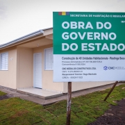 Uma fotografia colorida em plano m&eacute;dio e &acirc;ngulo frontal foca na placa oficial de obra p&uacute;blica instalada em frente a uma resid&ecirc;ncia rec&eacute;m-constru&iacute;da num loteamento habitacional.

**A Placa de Obra (em primeiro plano &agrave; direita):**

* No lado direito da imagem, sobressai uma grande placa retangular com topo em tom verde-escuro e base branca.


* No topo verde, destaca-se no cabe&ccedil;alho o texto **"SECRETARIA DE HABITA&Ccedil;&Atilde;O E REGULARIZA&Ccedil;&Atilde;O FUNDI&Aacute;RIA"** e, abaixo em letras garrafais brancas mai&uacute;sculas: **"OBRA DO GOVERNO DO ESTADO"**. No canto superior direito, l&ecirc;-se **"INVESTIMENTO TOTAL R$ 6.159.999,60"** e um QR Code.


* Na parte inferior branca, constam as informa&ccedil;&otilde;es t&eacute;cnicas e institucionais:


* **T&iacute;tulo:** *"Constru&ccedil;&atilde;o de 40 Unidades Habitacionais - Restinga S&ecirc;ca / RS"*.


* **Processo/Concorr&ecirc;ncia:** *"Processo Administrativo N.&ordm; 25/1700-0001725-0 | Concorr&ecirc;ncia 9192/2024"*.


* **Empresa e Respons&aacute;vel:** *"CMC M&Oacute;DULOS CONSTRUTIVOS LTDA. | Respons&aacute;vel T&eacute;cnico: Hugo Machado CREA - 5062411522"*.


* **Log&oacute;tipos:** A marca *"CMC MODULAR"* ao centro e o bras&atilde;o oficial do *"GOVERNO DO ESTADO RIO GRANDE DO SUL"* no canto inferior direito.





**A Casa e o Entorno (&agrave; esquerda e ao fundo):**

* &Agrave; esquerda, em segundo plano, v&ecirc;-se a fachada de uma casa t&eacute;rrea novinha em folha com paredes pintadas em tom bege-claro/areia e n&uacute;mero residencial **"1721"** fixado na parede.


* O im&oacute;vel possui porta de entrada e janelas com venezianas brancas fechadas, marquise com l&acirc;mpada acesa e calha prateada no beiral do telhado.


* &Agrave; frente da resid&ecirc;ncia, estende-se uma faixa de relva rec&eacute;m-plantada e caminho de acesso em concreto. Ao fundo, avista-se a continua&ccedil;&atilde;o da fileira de casas do conjunto habitacional sob um c&eacute;u nublado.