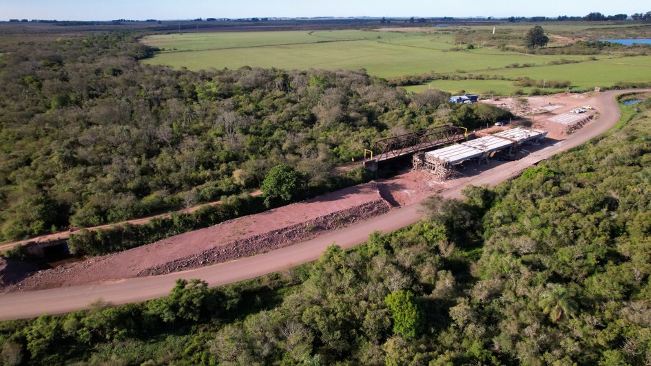 Uma fotografia a&eacute;rea colorida em plano geral e perspetiva obl&iacute;qua (capturada por drone) regista a vista panor&acirc;mica das obras de infraestrutura e reconstru&ccedil;&atilde;o de uma ponte rodovi&aacute;ria num ambiente rural e nativo.

**A Ponte e o Canteiro de Obras (ao centro e &agrave; direita):**

* No centro-direito da composi&ccedil;&atilde;o, observa-se a estrutura em constru&ccedil;&atilde;o de uma nova ponte de bet&atilde;o armado, com os tabuleiros e pilares de sustenta&ccedil;&atilde;o montados.


* Paralelamente &agrave; nova ponte, encontra-se a estrutura met&aacute;lica de uma antiga ponte treli&ccedil;ada de tom escuro.


* Ao lado das estruturas, estende-se uma via de terra batida e cascalho avermelhado. Na extremidade superior da curva, avista-se o canteiro de apoio com contentores, ve&iacute;culos e materiais de constru&ccedil;&atilde;o organizados.



**A Paisagem e a Vegeta&ccedil;&atilde;o:**

* A via e as pontes cruzam uma vasta &aacute;rea rodeada por uma densa mata nativa e &aacute;rvores de copa verde-escura.


* Ao fundo e em redor da &aacute;rea florestal, a paisagem abre-se em extensos campos de cultivo verdes e pastagens sob a linha do horizonte, sob ilumina&ccedil;&atilde;o solar direta e c&eacute;u claro num tom azul-suave.