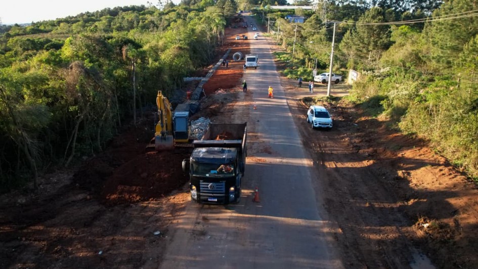 Foto a&eacute;rea mostra extens&atilde;o da estrada. Pr&oacute;ximo est&aacute; um caminh&atilde;o e retroescavadeira que trabalham nas margens. Nos dois lados da rodovia, h&aacute; mata fechada. 