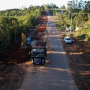 Foto a&eacute;rea mostra extens&atilde;o da estrada. Pr&oacute;ximo est&aacute; um caminh&atilde;o e retroescavadeira que trabalham nas margens. Nos dois lados da rodovia, h&aacute; mata fechada. 
