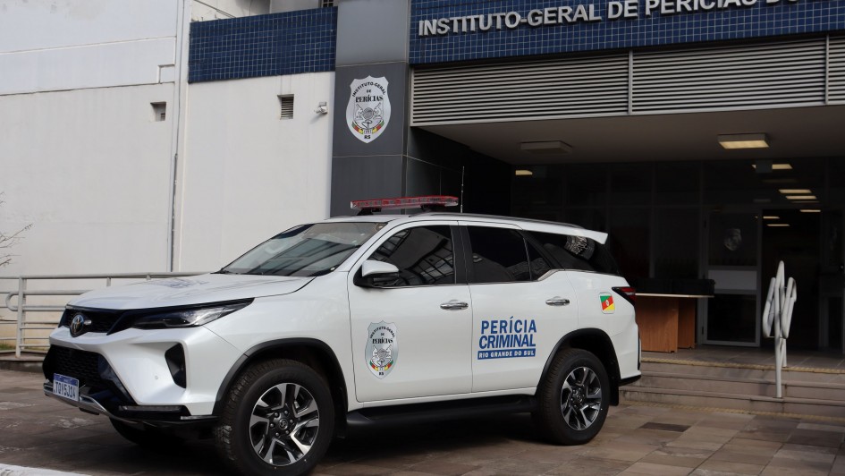 Uma fotografia colorida em plano m&eacute;dio e perspectiva em tr&ecirc;s quartos capta uma viatura oficial de Per&iacute;cia Criminal estacionada em frente &agrave; sede do Instituto-Geral de Per&iacute;cias do Rio Grande do Sul (IGP-RS).

**A Viatura Oficial (em primeiro plano):**

* No centro e &agrave; esquerda da imagem, destaca-se um ve&iacute;culo utilit&aacute;rio desportivo (SUV) oficial da marca Toyota, de cor branca, visto de perfil diagonal de tr&ecirc;s quartos.
* Na porta lateral do condutor, v&ecirc;-se o bras&atilde;o do **"INSTITUTO-GERAL DE PER&Iacute;CIAS RS"** e a inscri&ccedil;&atilde;o em letras azuis e pretas: **"PER&Iacute;CIA CRIMINAL - RIO GRANDE DO SUL"**.
* Junto &agrave; janela traseira, observa-se a bandeira do Estado do Rio Grande do Sul. No teto do ve&iacute;culo, encontra-se instalado um sinalizador luminoso (giroflex) de emerg&ecirc;ncia em tom vermelho-escuro.

 O Edif&iacute;cio do IGP (ao fundo e &agrave; direita):

* Ao fundo, ergue-se a fachada do pr&eacute;dio oficial. Na parte superior da entrada, sobre uma faixa de azulejos ou pastilhas azuis, sobressai a inscri&ccedil;&atilde;o em letras garrafais brancas mai&uacute;sculas: **"INSTITUTO-GERAL DE PER&Iacute;CIAS DO RI..."**
* Num pilar cinzento &agrave; esquerda da entrada principal, est&aacute; fixado o bras&atilde;o oficial do Instituto-Geral de Per&iacute;cias.
* &Agrave; direita, avista-se a entrada do edif&iacute;cio com portas de vidro, escadaria de acesso com corrim&atilde;o met&aacute;lico prateado e rampas. O piso exterior &eacute; feito de lajotas de pedra sob ilumina&ccedil;&atilde;o natural diurna.