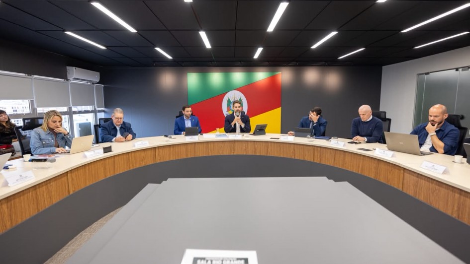Uma fotografia colorida em plano geral e perspetiva em &acirc;ngulo levemente baixo capta o governador Eduardo Leite reunido com secret&aacute;rios de Estado ao redor de uma grande mesa semicircular de reuni&otilde;es numa sala institucional.

**A Mesa Principal e as Autoridades (ao centro e em primeiro plano):**

* Ao centro da mesa semicircular em tom de madeira clara, o governador Eduardo Leite fala ao microfone com as m&atilde;os entrela&ccedil;adas. Ele veste blazer escuro sobre camisa clara.


* Ao redor da mesa, dispostos em curva, secret&aacute;rios e secret&aacute;rias acompanham a reuni&atilde;o com computadores port&aacute;teis, tablets e blocos de anota&ccedil;&otilde;es sobre a bancada, al&eacute;m de identificadores nominais &agrave; frente de cada lugar.


* &Agrave; esquerda do governador, v&ecirc;-se um homem de blazer azul e, na extremidade esquerda, uma mulher de casaco de ganga a trabalhar num port&aacute;til.


* &Agrave; direita do governador, encontram-se outros quatro secret&aacute;rios e assessores em trajes formais e esporte fino (blazers e camisas sociais), observando a apresenta&ccedil;&atilde;o ou consultando os seus dispositivos eletr&oacute;nicos.




* Em primeiro plano, no centro da composi&ccedil;&atilde;o, sobressai o tampo cinzento de um m&oacute;dulo da mesa com uma folha impressa onde se l&ecirc; **"SALA RIO GRANDE"**.



**O Ambiente e a S&iacute;mbolo Oficial (ao fundo):**

* Na parede do fundo, em posi&ccedil;&atilde;o central de destaque atr&aacute;s do governador, sobressai um grande painel retangular com a bandeira do Estado do Rio Grande do Sul e o seu bras&atilde;o de armas impresso no centro.


* A sala possui paredes escuras e teto preto com linhas paralelas de ilumina&ccedil;&atilde;o LED encastradas e acesas, garantindo uma luz clara e focada sobre a mesa de reuni&otilde;es. &Agrave; esquerda, avistam-se janelas com persianas cinzentas.