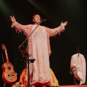 Foto mostra a cantora Adriana Calcanhotto no palco. Ela est&aacute; em p&eacute;, em frente ao microfone. Ela est&aacute; cantando com os bra&ccedil;os abertos. Ela usa uma roupa em tonalidade bege. Atr&aacute;s dela, h&aacute; um viol&atilde;o num pedestal. O fundo &eacute; todo preto. 