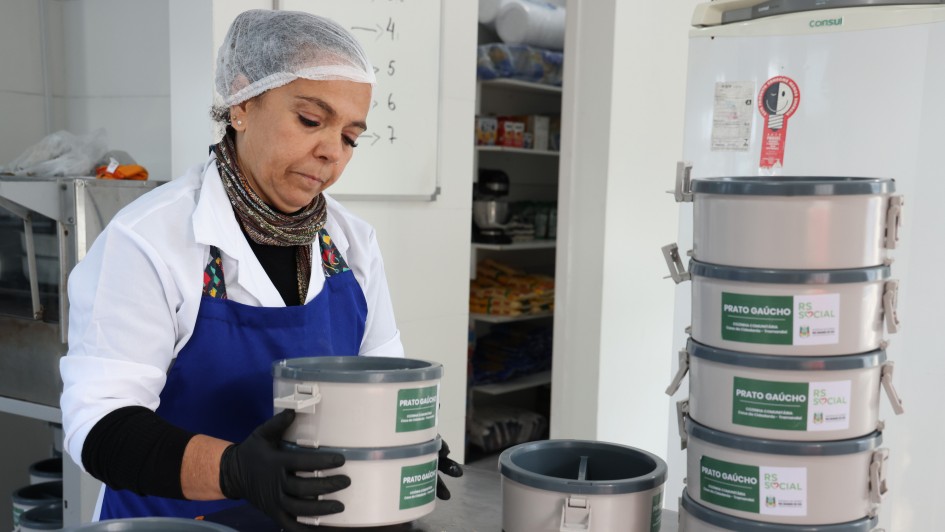 A imagem mostra uma mulher com avental e touca manuseando uma vianda com o nome do programa "Prato Ga&uacute;cho". Ao lado, em cima de uma bancada, h&aacute; uma pilha de viandas retorn&aacute;veis. 