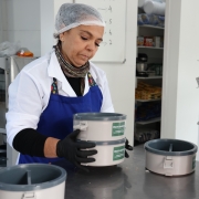 A imagem mostra uma mulher com avental e touca manuseando uma vianda com o nome do programa "Prato Ga&uacute;cho". Ao lado, em cima de uma bancada, h&aacute; uma pilha de viandas retorn&aacute;veis. 