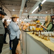 A imagem mostra um estande em um pavilh&atilde;o com produtos da agricultura familiar. Um casal observa os produtos e uma vendedora est&aacute; posicionada atr&aacute;s do balc&atilde;o. 